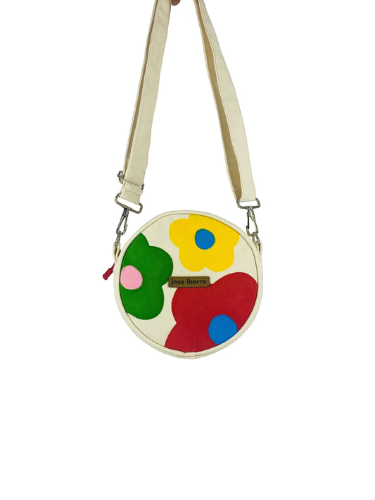 Jose Ibarra Estudio cream canvas round crossbody bag with hand-painted red, green, yellow, and blue flowers. Includes adjustable strap.