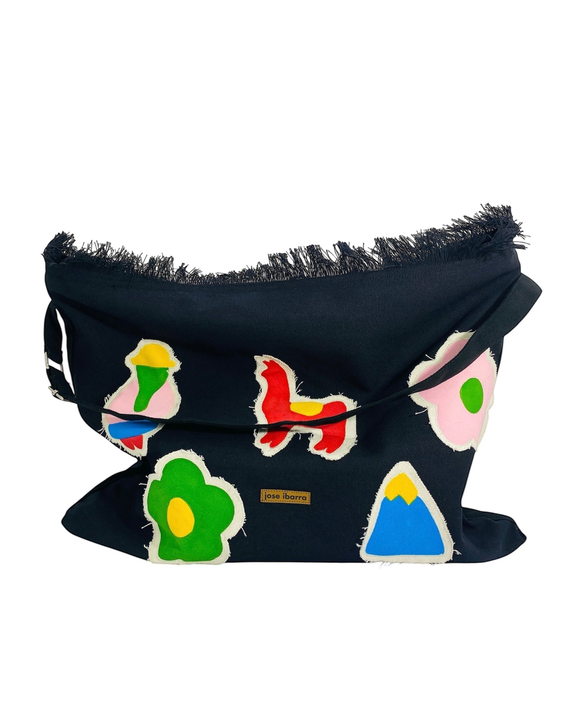 Jose Ibarra black cotton canvas tote bag with colorful hand-painted patches of flowers, mountains, and abstract figures. Features frayed edges and an adjustable strap.