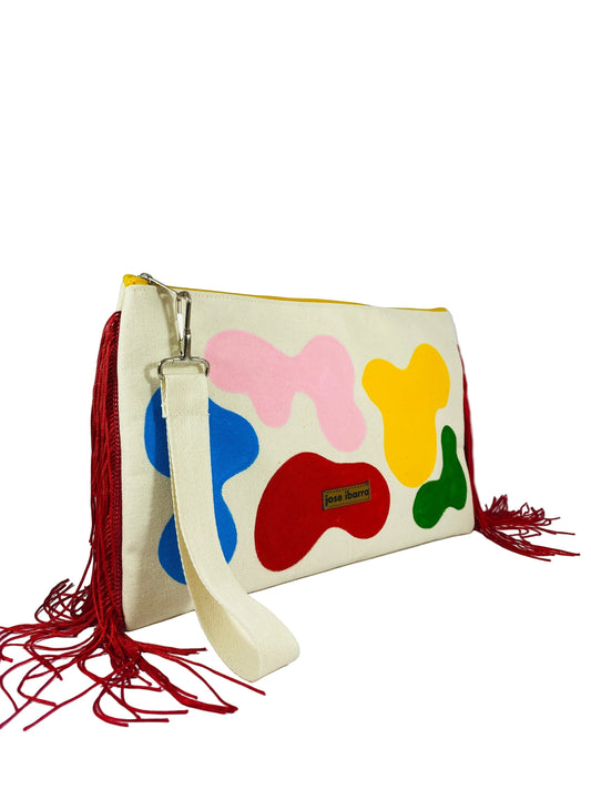 Jose Ibarra Estudio large cream cotton clutch with hand-painted abstract shapes in red, blue, pink, yellow, and green. Features red fringe details and a cream wrist strap.