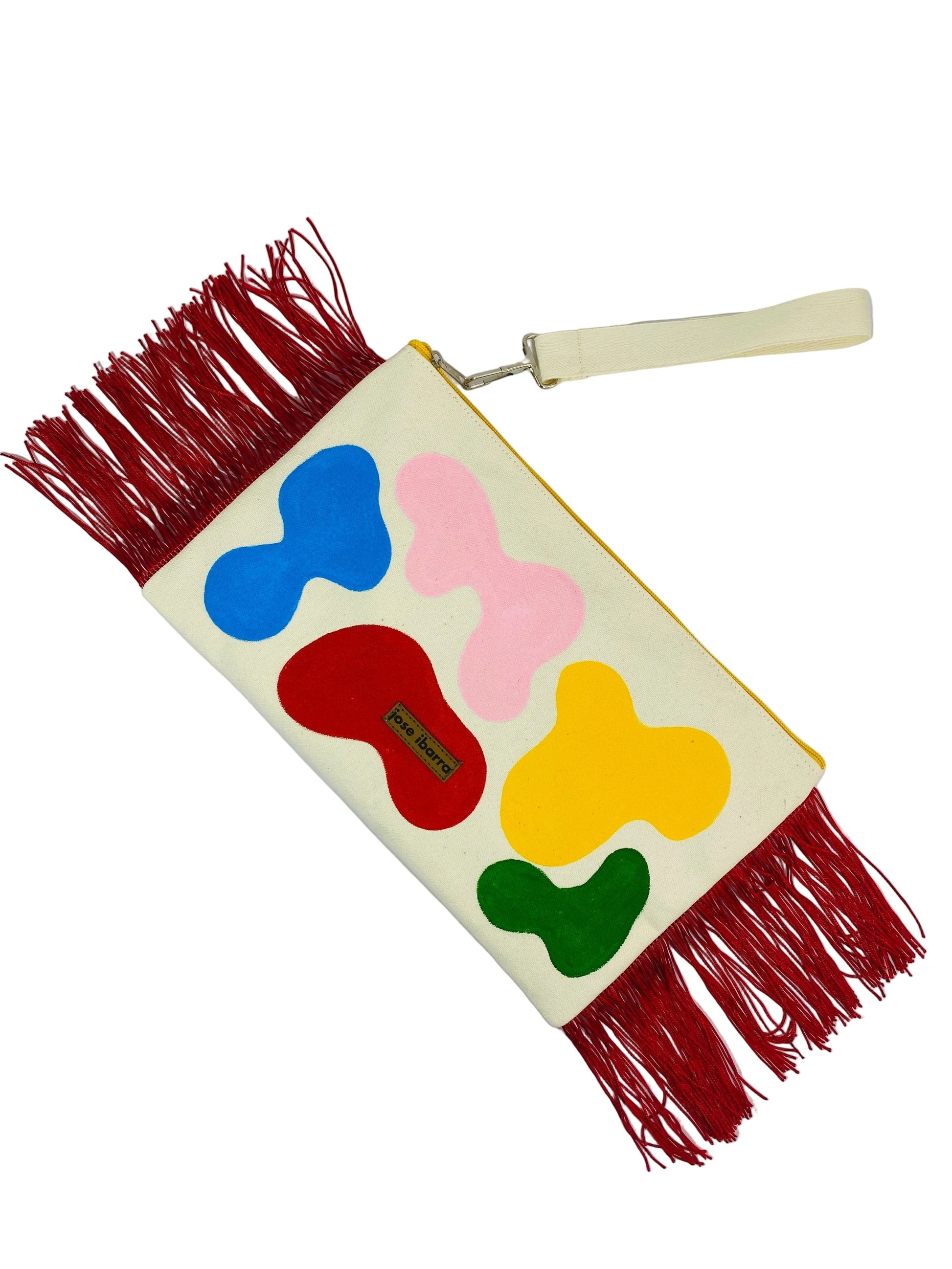 Jose Ibarra Estudio large cream cotton clutch with hand-painted abstract shapes in red, blue, pink, yellow, and green, and red fringe detail.