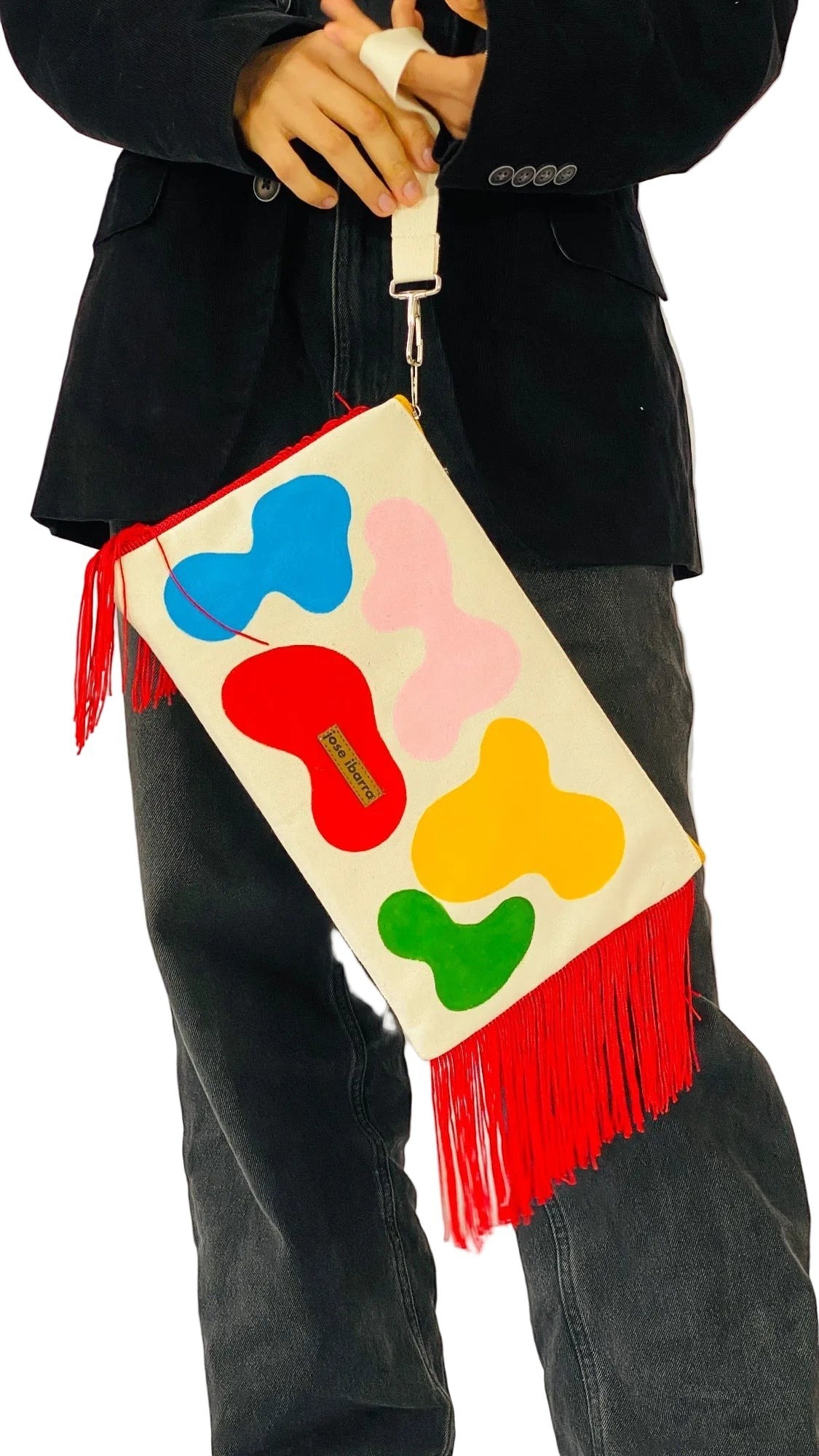 Jose Ibarra Estudio large cream cotton clutch with hand-painted abstract shapes in red, blue, pink, yellow, and green, featuring red fringe and a wrist strap.