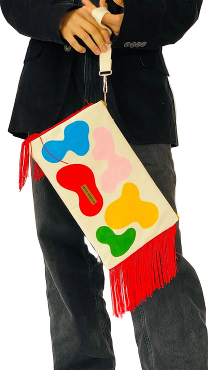 Jose Ibarra Estudio large cream cotton clutch with hand-painted abstract shapes in red, blue, pink, yellow, and green, featuring red fringe and a wrist strap.