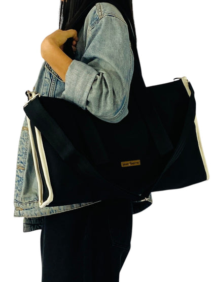 Jose Ibarra Estudio black canvas tote bag with white trim and adjustable strap, worn by a person in a denim jacket.