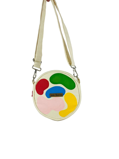 Jose Ibarra Estudio cream canvas round crossbody bag with hand-painted abstract shapes in red, blue, green, pink, and yellow. Includes strap.