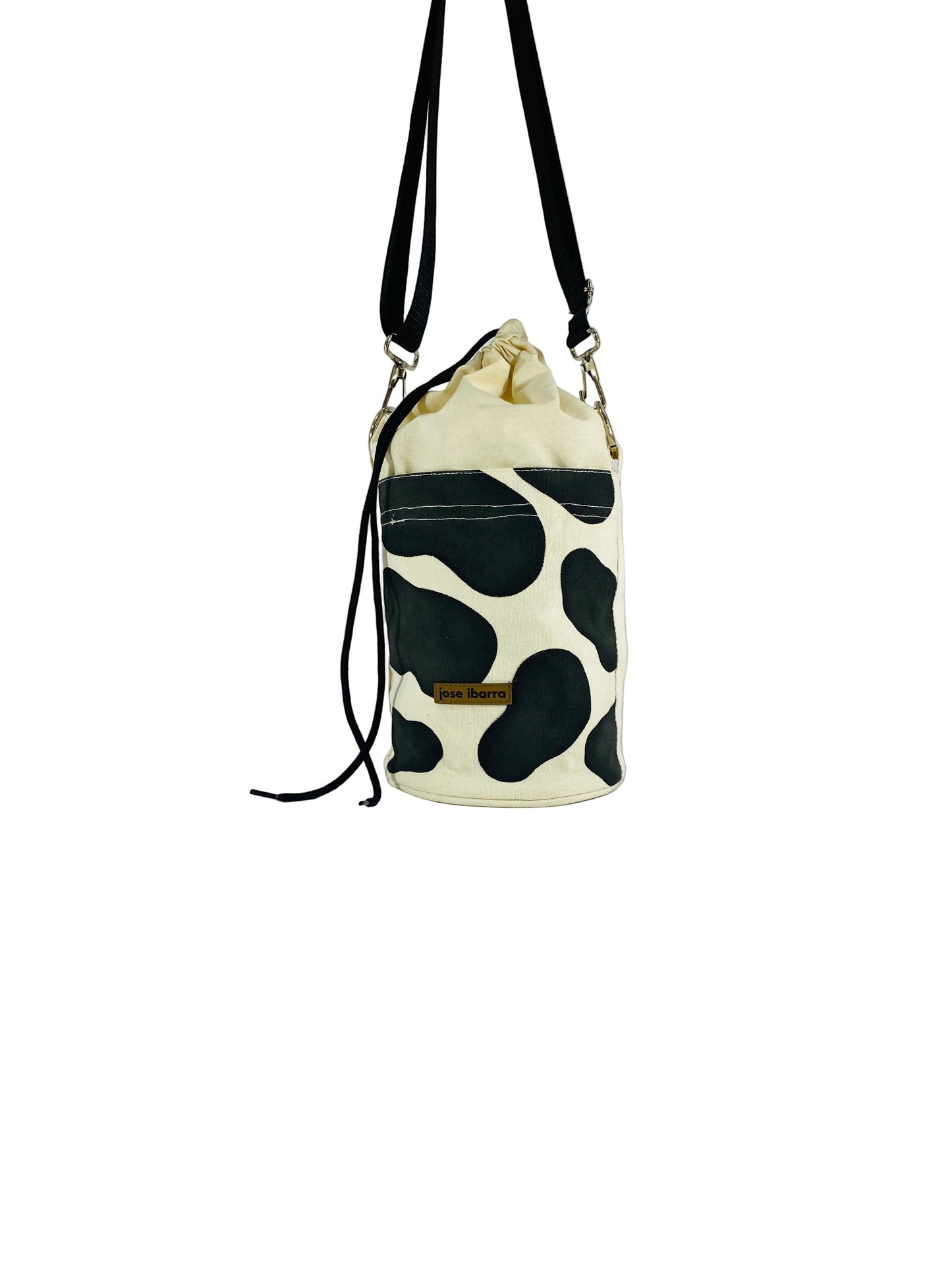 Jose Ibarra Estudio cream canvas bucket bag with black cow print, drawstring closure, and adjustable strap.