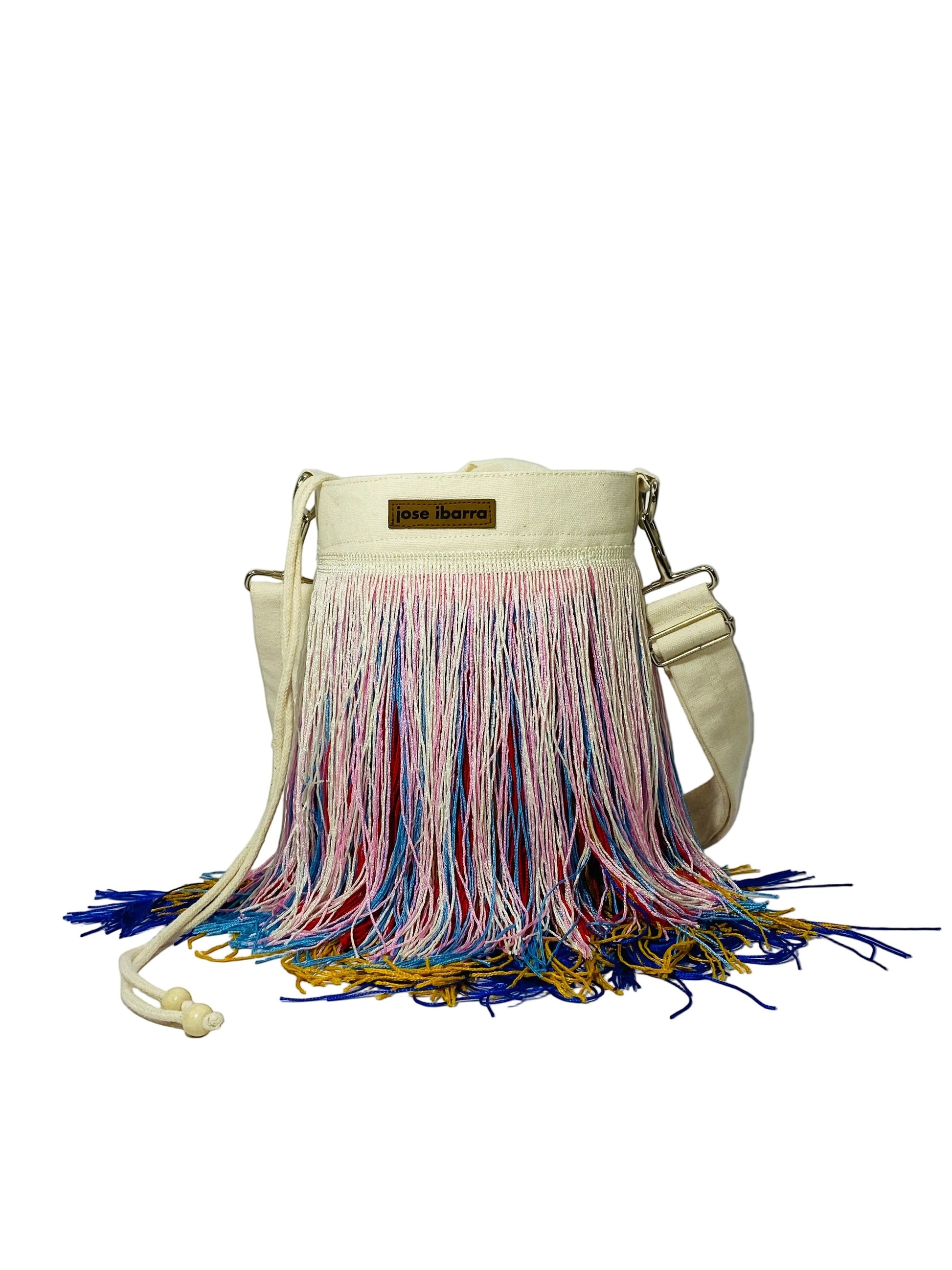 Jose Ibarra Estudio cream canvas bucket bag with multicolor fringe detailing and adjustable strap. Hand-painted, artisanal design.